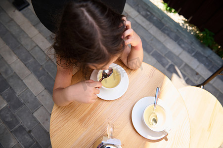 童男童女摄影照片_一个可爱的孩子坐在夏季露台的桌子旁，用勺子喝着健康的草药饮料的顶视图。