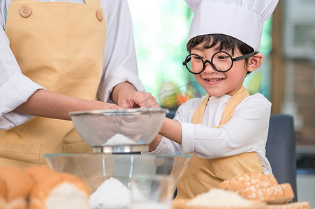 可爱的亚洲小男孩和美丽的母亲在家庭厨房的桌子上用筛子滤锅筛面粉，准备烘烤面包店和蛋糕。