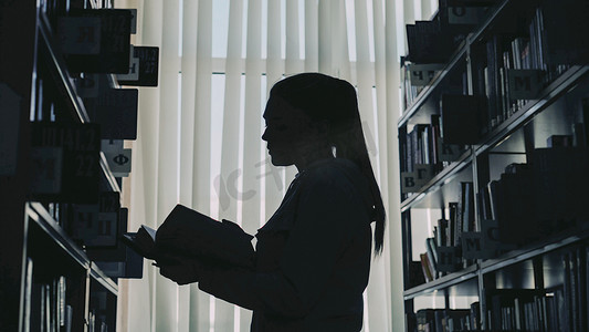 书翻页摄影照片_年轻女学生站在大图书馆书架间靠窗拿着书翻页阅读的剪影