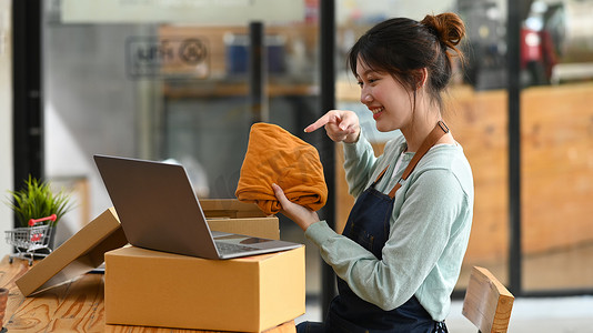 金融直播摄影照片_年轻的女性小企业主在笔记本电脑上在线直播时展示衣服。
