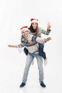 骑行背景摄影照片_圣诞节概念 — 身穿毛衣的年轻幸福夫妇享受白色灰色背景中的背驮式骑行