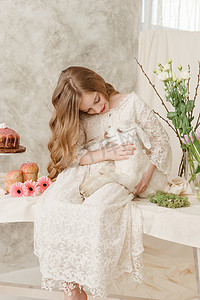 这个女孩坐在复活节餐桌旁，手里拿着蛋糕、春天的花朵和鹌鹑蛋，怀里抱着一个白色的孩子。