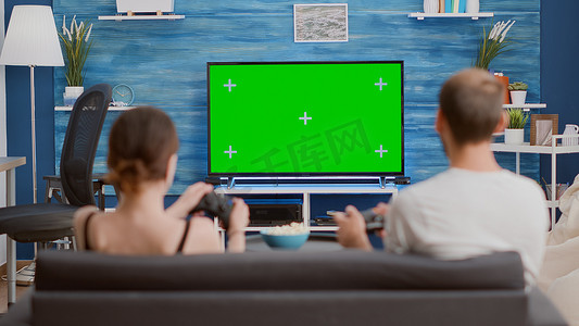 年轻女性和男朋友游戏玩家拿着控制器在绿屏电视上的控制台上玩动作游戏的后视图