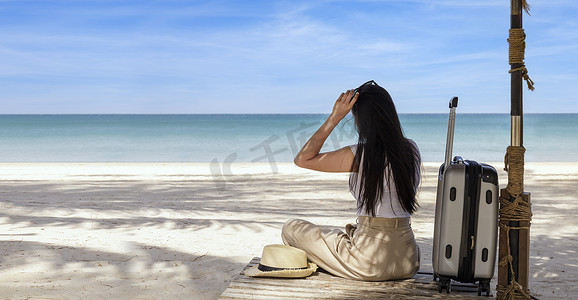 旅行箱旅游摄影照片_夏天，年轻快乐的女人带着旅行箱坐在海滩上。