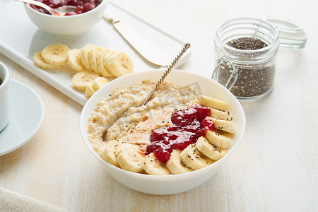 浅色摄影照片_燕麦粥、健康素食早餐的特写，包括草莓酱、花生酱、香蕉、白色木质浅色背景的奇亚籽。