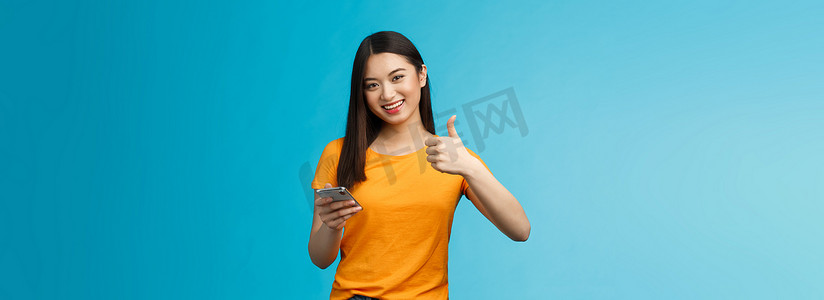满意的可爱亚洲黑发持有智能手机竖起大拇指很高兴，推荐很棒的小工具，批准好的新应用程序，同意朋友发布好的博客文章，站在蓝色背景