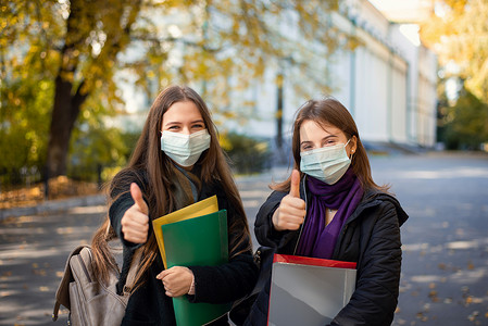 两名戴着医用防护口罩的女学生站在大学校园里对着镜头竖起大拇指