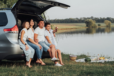 十几岁的青少年摄影照片_快乐的四名白人家庭成员坐在小型货车的后备箱里，父母带着两个十几岁的孩子、儿子和女儿在湖边户外周末野餐