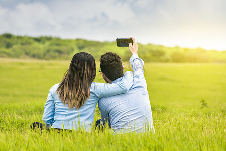 相爱的摄影照片_相爱的年轻情侣在田野里自拍，相爱的人们用智能手机在田野里自拍，微笑的相爱情侣坐在草地上自拍