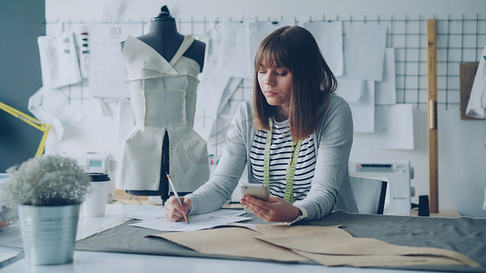 创意服装设计师在现代裁缝店的缝纫台工作时，正在查看智能手机并绘制草图。