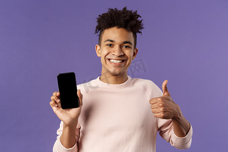手机批准摄影照片_快乐、满意的年轻男性顾客的特写肖像建议购买游戏或应用程序订阅，通过应用程序手机学习语言，竖起大拇指表示批准