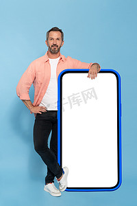 健康的中年男子靠在巨大的智能手机上，智能手机的白色屏幕是蓝色的，他穿着桃色衬衫和蓝色背景的黑色牛仔裤，在镜头前开心地微笑着。