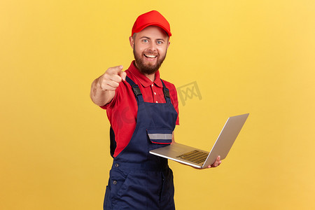 穿着蓝色工作服、红色 T 恤和帽子的工人在笔记本电脑上工作，手指指向相机。