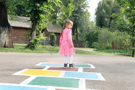 跳房子摄影照片_穿着粉色裙子的小女孩在户外操场上玩跳房子，儿童户外活动