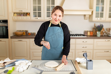现代厨房里，穿着围裙的亚洲女厨师看着相机，在硅胶烤垫上的煮熟的小麦糕点面团上撒上面粉
