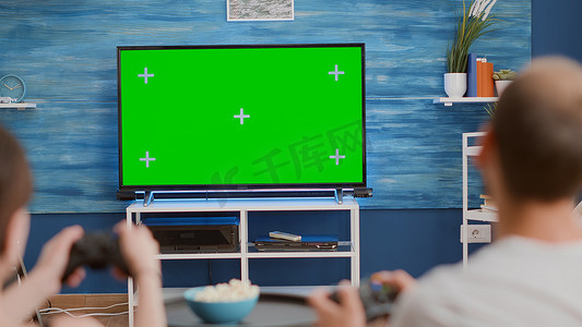 游戏钥匙摄影照片_一对夫妇拿着控制器坐在沙发上的绿屏电视上玩肌动蛋白控制台游戏的特写