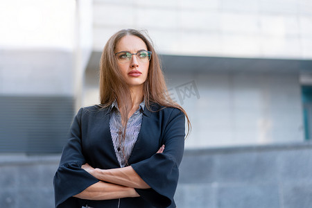 长发、身穿黑色夹克的女商人站在公司办公楼附近的户外，双手交叉