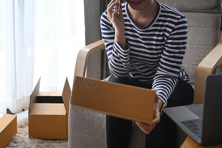 年轻女子通过智能手机确认顾客订单，并准备包裹箱以便在家庭办公室送货。
