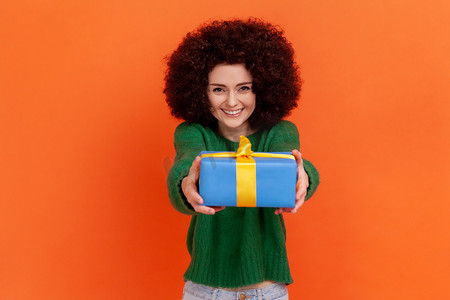 迷人的非洲发型女人的肖像，穿着绿色休闲风格的毛衣送礼物，拿着蓝色包装的礼品盒。