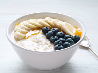 燕麦片配香蕉、蓝莓、奇亚籽、果酱、蜂蜜，白色木质背景。
