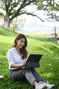 有魅力的年轻女子坐在公园的草地上，用电脑平板电脑在线工作。