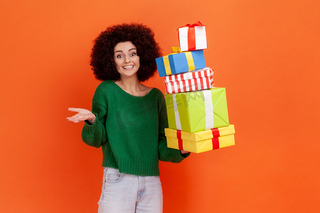 绿色购物节摄影照片_微笑的非洲发型女人穿着绿色休闲风格的毛衣，拿着礼物盒，把手伸到一边，表达积极的情绪。
