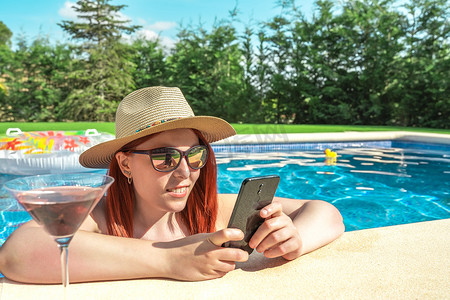 年轻的红发女子在泳池边放松，喝着鸡尾酒，看着她的智能手机。