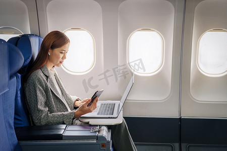 一位成功的亚洲女商人或女性企业家在飞机上穿着正装，坐在商务舱座位上，在飞行过程中使用带有笔记本电脑的文件