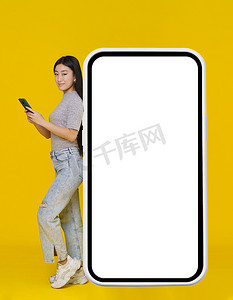 手里拿着手机、微笑的亚洲女孩靠在巨大的智能手机、带有白色、黑色屏幕的数字平板电脑上，穿着黄色背景的休闲装。