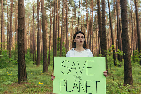 保护环境海报摄影照片_年轻女活动家站在树林里，举着拯救地球的海报，一名志愿者与森林砍伐作斗争