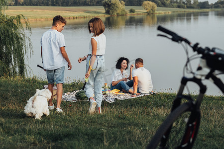 中年男女野餐，两个十几岁的孩子、儿子和女儿与宠物狗玩耍，幸福的四口之家在湖边享受户外周末