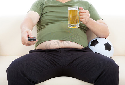 电视摄影照片_胖子喝啤酒坐在沙发上看电视