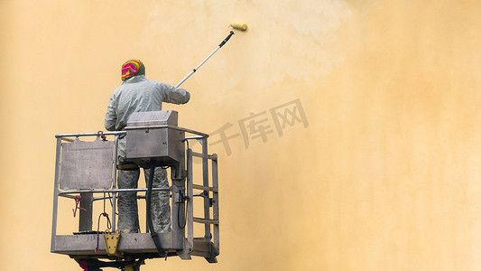 电梯平台上的人在室外用滚子涂墙。建筑工地梯子上的工人手绘黄墙