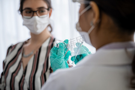 戴口罩、手套和面罩的女医生或护士拿着注射器和验尸犬19疫苗，一名女性坐在实验室等待注射疫苗。预防COVID-19的概念.