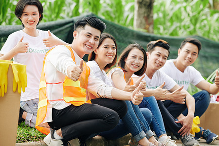 快乐的亚洲学生志愿者们坐在户外，微笑着，竖起大拇指