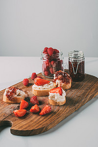 草莓果酱摄影照片_关闭拍摄的切割板与草莓果酱在罐子和三明治奶油奶酪和果酱灰色 