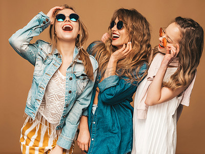 三个年轻美丽的微笑的嬉皮士女孩在时尚夏季休闲牛仔裤礼服。性感无忧无虑的女人摆在金色的墙附近。积极的模特疯狂的太阳镜