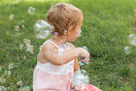 Side view of baby girl in dress holding soap bubbles in summer park 