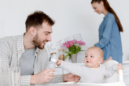 微笑的父亲喂养婴儿奶瓶牛奶, 母亲站在厨房后面
