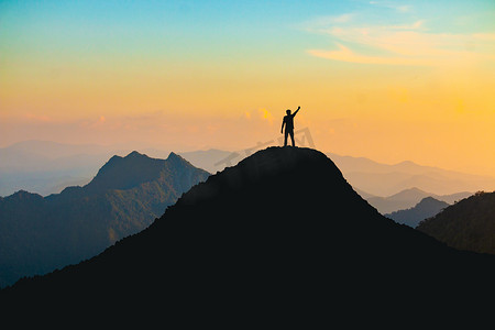 轮廓摄影照片_在日落时分，一个手拿着愉快的高举在山顶上的人的轮廓：努力与成功的概念