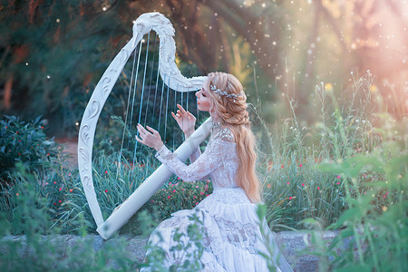 礼服首饰摄影照片_神秘的森林仙女在神话般的地方玩白色竖琴，女孩与长长的金发和优雅的蕾丝复古礼服呼吁明亮的阳光，夫人与银首饰和乐器