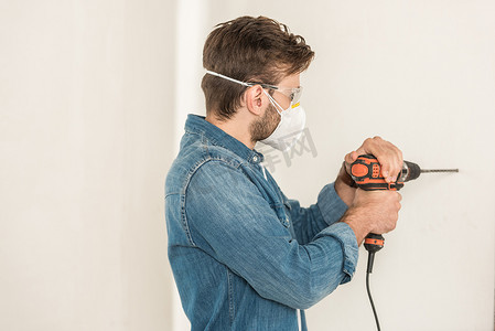 在房屋维修过程中使用电钻在墙上使用电钻的年轻人穿着防护工作服 