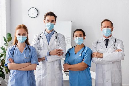 多文化医生和戴口罩的护士，双手交叉，站在医院里看着相机