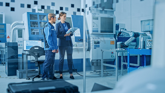 工厂车间：专业的女工程师和男机械操作员使用工业数字制表计算机编程机器人臂生产线。配备数控机床的高科技设施