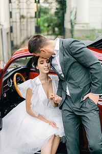 新娘和新郎在热带花园一辆红色复古汽车的背景下接吻.