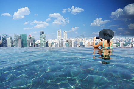 2018摄影照片_美丽的女人与黑色太阳帽钉图片与智能手机到新加坡金融区和天际线从滨海湾金沙无限游泳池，新加坡，2018年10月12日