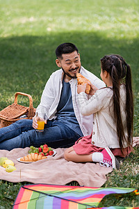 Girl holding croissant near cheerful asian dad with orange juice on blanket in park 