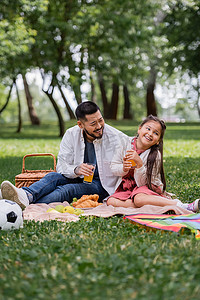 Happy asian family holding orange juice near food and football in summer park 