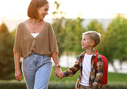 妈妈和小儿子，背着背包上学，在阳光灿烂的秋天里一起穿过公园上学，可爱快乐的男孩微笑着看着妈妈，准备学习