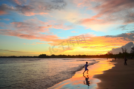 反映摄影照片_海洋海岸上的热带日落。这孩子在海滩上跳来跳去。明亮的天空颜色反映在水和沙子中。斯里兰卡